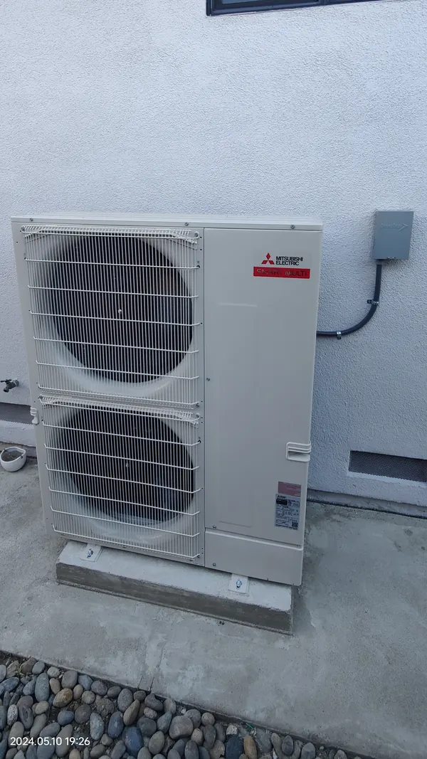 A Mitsubishi Electric SMART MULTI outdoor unit with twin fans on a concrete pad against a stucco wall, a grey disconnect box above it
