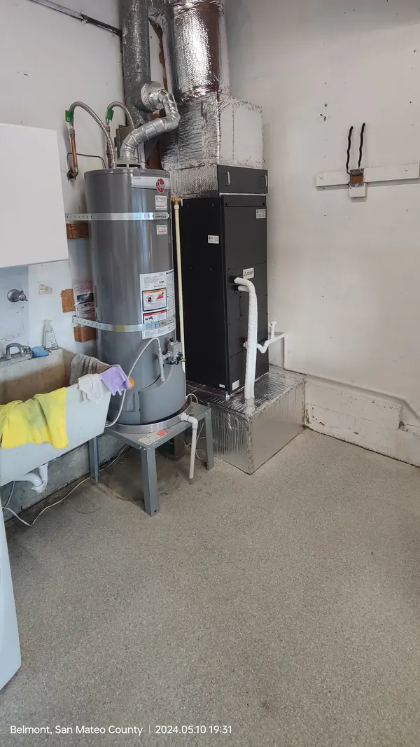 A black air handler on a foil-wrapped stand beside a water heater and a utility sink in a garage, insulated duct above it