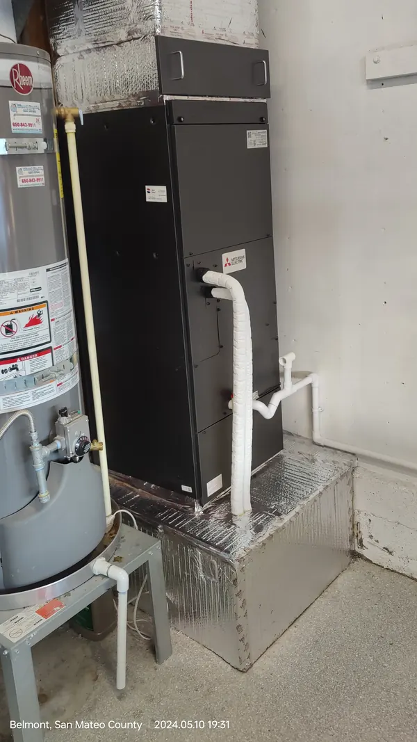 A close view of the black air handler on its foil-wrapped stand with white pipes running down its side