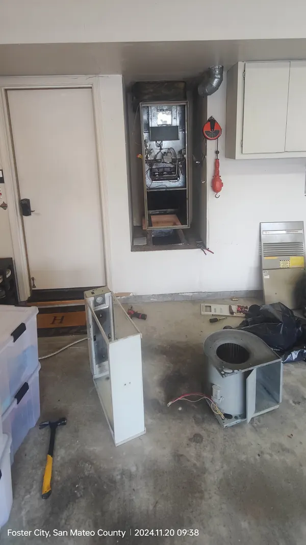 An old furnace opened up in a garage wall alcove, its blower and cabinet panels on the concrete floor in front of it