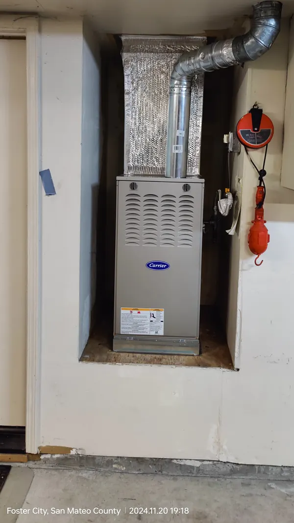 A new grey Carrier furnace in a garage wall alcove with a foil-wrapped plenum and a metal vent pipe above it