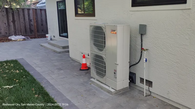 A Mitsubishi Electric SMART MULTI outdoor unit on a pad beside a stucco house, with a paved path, a lawn and an orange safety cone