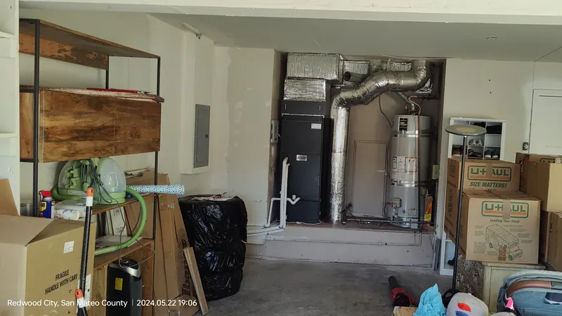 A garage with shelves and stacked boxes, and at the back a black air handler beside a water heater with foil-wrapped duct above