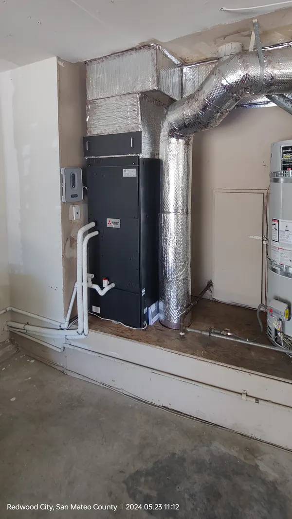 A close view of a black air handler on a wooden platform with foil-wrapped duct and white pipes running down to the floor