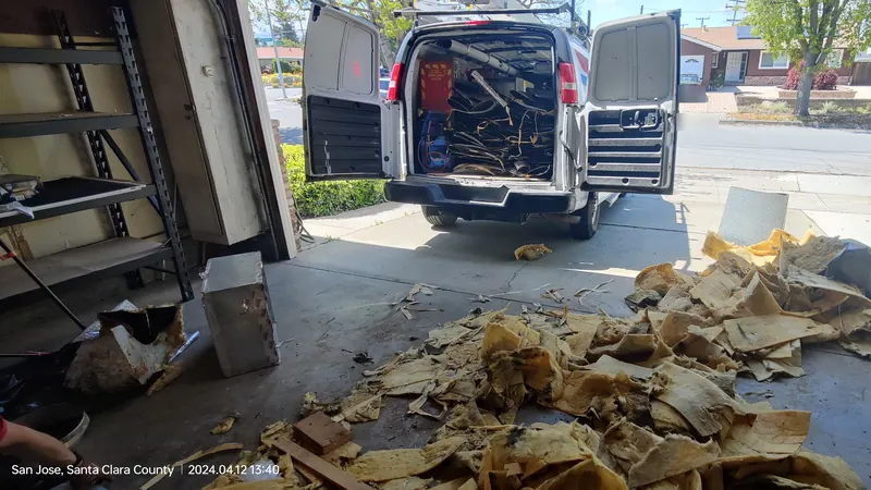 A work van with its back doors open and old duct loaded inside, with torn insulation piled on the driveway in front of the garage