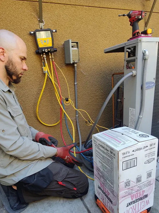 A technician kneels with manifold gauges and a refrigerant cylinder beside an outdoor unit