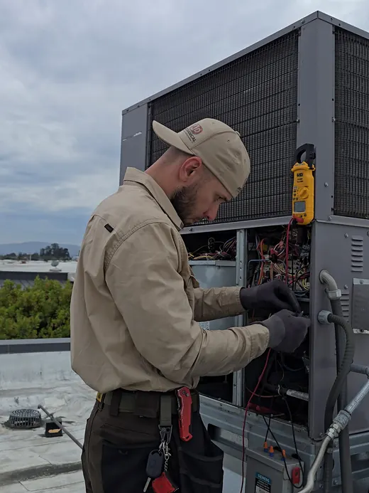 A technician reads a clamp meter at a rooftop unit's wiring