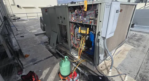 An open rooftop package unit with gauges hooked up and a refrigerant cylinder beside it